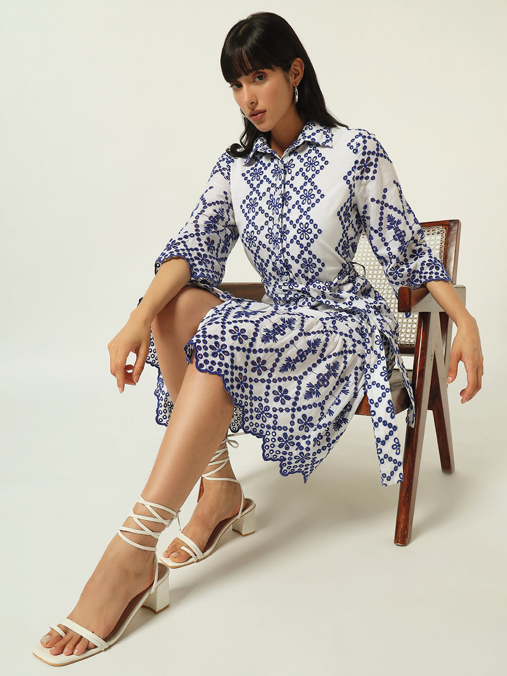 A woman seated on a chair wearing a knee-length blue and white geometric patterned dress with a spread collar and 3/4th sleeves, paired with white strappy sandals.
